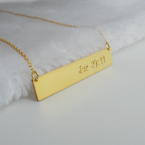 May include: Gold bar necklace with the inscription "Jer 29:11" hanging on a white fuzzy surface.