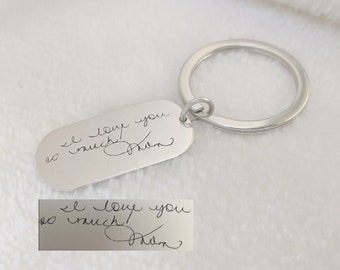 Könnte beinhalten: Silberner Schlüsselanhänger mit einem rechteckigen Anhänger, der mit der handschriftlichen Nachricht "Ich liebe dich so sehr!" und einer Unterschrift graviert ist.