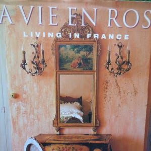 May include: A book cover titled LA VIE EN ROSE LIVING IN FRANCE by Suzanne Lowry with photographs by Tim Clinch. The cover features a vanity table with a floral painted finish, a matching chair, a wall mirror, and two wall-mounted candle sconces.
