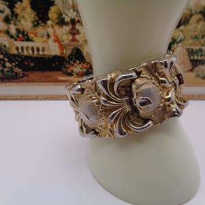 May include: A wide cuff bracelet featuring a two-tone design with alternating gold and silver-colored metal sections. The surface displays intricate embossed floral and scrollwork patterns with textured circular elements throughout the piece.