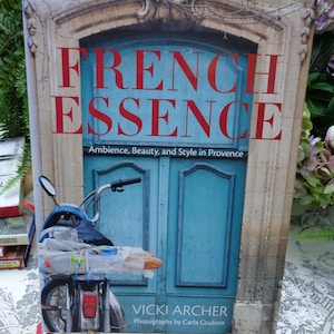 May include: A hardcover book titled French Essence: Ambience, Beauty, and Style in Provence by Vicki Archer with photographs by Carla Coulson. The cover features a blue door and a blue moped carrying a baguette in a plastic bag.
