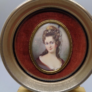 May include: A gold-framed oval portrait of a woman with a white head scarf and a pearl necklace. The portrait is set against a red velvet background.