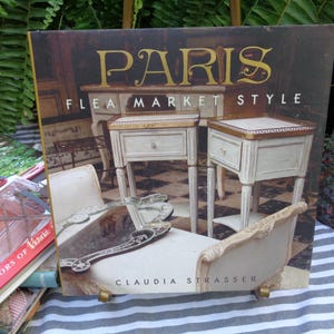 May include: A hardcover book titled Paris Flea Market Style by Claudia Strasser. The cover features a photograph of two off-white bedside tables with gold-toned trim, a chaise lounge, and a decorative mirror. The text is printed in gold and white.