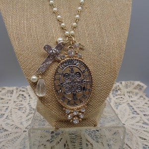 May include: A gold-toned necklace with a pendant featuring an oval medallion with religious text and a cross. The necklace has a pearl chain and is adorned with clear crystals and a teardrop-shaped crystal.