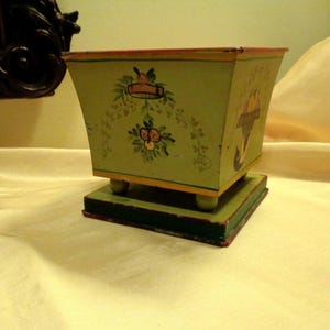 May include: A vintage, hand-painted wooden box with a light green exterior. The box features floral and banner designs, with yellow and red accents. It sits on a green base with small, round feet, showcasing a rustic aesthetic.
