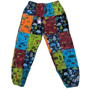 Op de afbeelding: Patchworkbroek met een kleurrijk paddenstoelenpatroon. De broek heeft een taille met trekkoord en is gemaakt van verschillende stoffen vierkanten in tinten blauw, groen, oranje en bordeauxrood. De paddenstoelontwerpen variëren in grootte en kleur, wat een levendige en unieke uitstraling creëert.