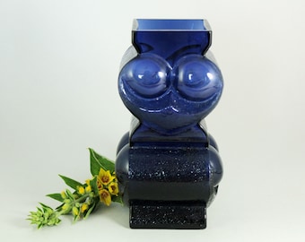 May include: A cobalt blue glass vase with a unique, face-like design. The vase has a square top and a rounded base with a textured surface.