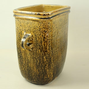 Op de afbeelding: Een getextureerde, rechthoekige vaas met een handvat. De vaas is voornamelijk bruin met zwarte accenten en een vierkante bovenkant. Het handvat is donkerbruin van kleur. De vaas is ongeveer 20 cm hoog.