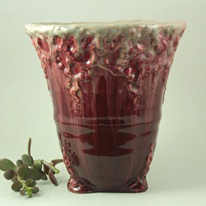 May include: A tall, burgundy-colored ceramic vase with a textured, organic design. The top rim has a light gray and white glaze, and the vase features a flared shape. A small succulent plant is visible to the left of the vase.