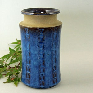 Puede incluir: Un jarrón de cerámica de forma cilíndrica, con un esmalte azul texturizado y detalles marrones verticales. El jarrón tiene un borde beige y una parte superior marrón oscuro. Artículo de decoración del hogar.
