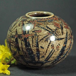 May include: A spherical ceramic vase with a beige base and a pattern of black and rust-coloured lines and shapes. The vase has a small opening and a light beige rim. The vase is a decorative item.