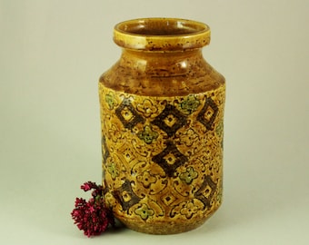 Könnte beinhalten: Eine braune Keramikvase mit einem geometrischen Muster in Braun-, Grün- und Gelbtönen. Die Vase hat eine strukturierte Oberfläche und eine leicht ausgestellte Öffnung.
