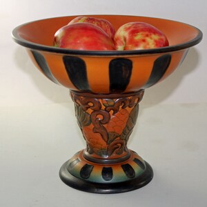 May include: A decorative orange and black striped fruit bowl with a pedestal base. The bowl contains several red apples. The pedestal features ornate carvings and a dark base, creating a striking contrast.
