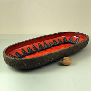 May include: A long, oval ceramic dish with a rough, textured exterior. The interior is glazed in a bright orange color with a black and blue geometric design.