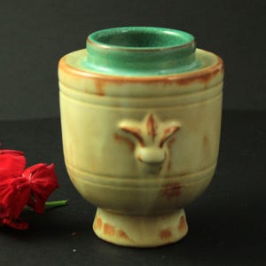 Może przedstawiać: Ceramiczny wazon z jasnożółtym korpusem i zieloną obwódką. Wazon posiada motyw fleur-de-lis i podstawę na cokole. Wazon ma wysokość około 13 cm.