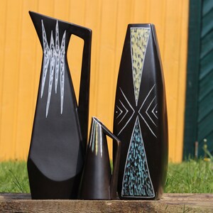 May include: Three black ceramic vases with geometric designs in white, yellow, and blue. The vases are arranged in a row on a wooden surface.