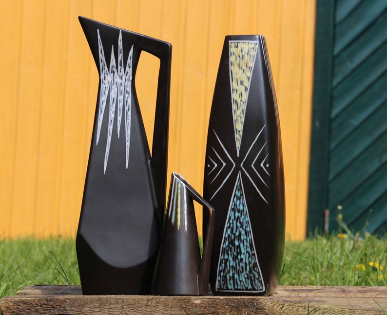 May include: Three black ceramic vases with geometric designs in white, yellow, and blue. The vases are arranged in a row on a wooden surface.