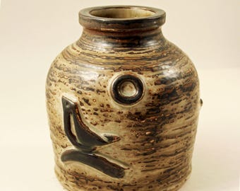 May include: A ceramic vase with a textured, speckled surface in shades of brown and tan. The vase features a dark brown, stylized bird design and a circular accent. The rim and top are a darker brown.