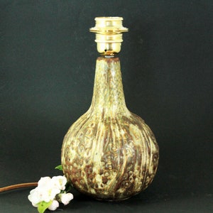 Könnte beinhalten: Eine Keramik-Tischlampe mit einer Lampenfassung. Die Lampe hat einen bauchigen Sockel mit einem strukturierten, gesprenkelten Muster in Braun-, Beige- und Grüntönen. Die Lampe ist etwa 25 cm hoch. Ein kleiner Strauß weißer Blumen liegt in der Nähe des Sockels.