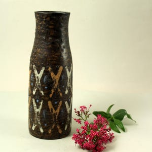 Pode incluir: Um vaso cilíndrico alto de cerâmica com uma superfície castanha texturizada. O exterior apresenta um padrão repetitivo de marcas em forma de X brancas e bronzeadas, intercaladas com pequenos pontos azul-claros numa grelha vertical.