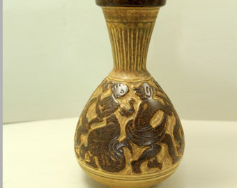 Peut inclure: Un vase en céramique aux tons bruns et beiges. Le vase présente un corps bulbeux avec un relief détaillé de personnages et un col cannelé. Le bord est brun foncé.