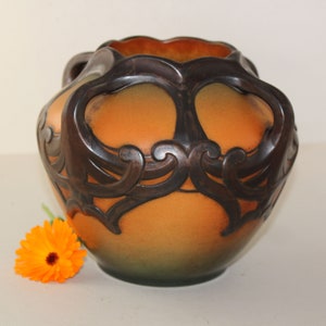 May include: A large, orange ceramic vase with intricate brown, raised, floral designs. The vase has a rounded body and two handles.