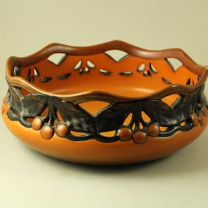 Puede incluir: Cuenco de cerámica naranja con un borde decorativo con diseño calado. El borde está adornado con hojas marrones oscuras y cerezas de color naranja-marrón. El cuenco tiene una forma lisa y redondeada.