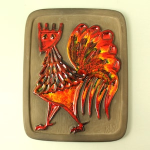 Puede incluir: Placa de pared rectangular de cerámica con un diseño de gallo estilizado. El gallo es principalmente rojo y naranja con detalles amarillos, sobre un fondo marrón. La obra de arte tiene un acabado texturizado y esmaltado.
