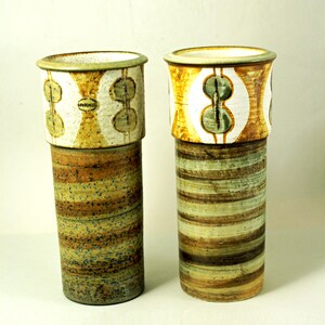 May include: Two ceramic vases with cylindrical bodies and flared rims. The upper sections feature a white background with a repeating pattern of green and yellow shapes. The lower sections have horizontal bands of brown, green, and tan.