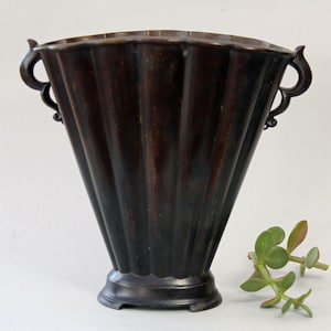 May include: A dark brown, fluted vase with two curved handles. The vase has a wide, fan-like shape and a scalloped rim. A small sprig of green leaves rests beside the vase.