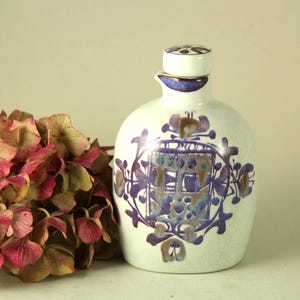 May include: A white ceramic bottle with a decorative blue and brown floral design. The bottle has a matching lid and a small spout. A bouquet of pink and brown flowers is next to the bottle.