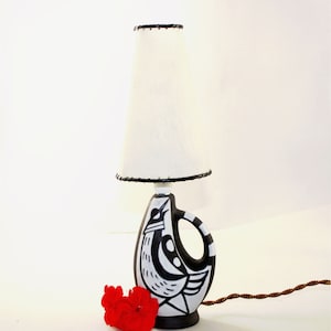 May include: A table lamp featuring a white lampshade and a black and white patterned base. The base has a handle and a geometric design. A red flower is placed near the base of the lamp.
