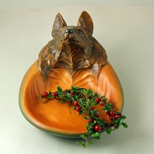May include: A wooden decorative bowl in the shape of a dog's head and body. The bowl is painted in shades of brown and orange, with a green rim. A sprig of red berries and green leaves rests inside the bowl.