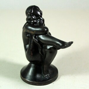 Può includere: Una scultura in bronzo scuro di una figura seduta, con le ginocchia sollevate, le braccia avvolte attorno alle gambe. La figura è seduta su una base rotonda. La scultura ha una finitura liscia e lucida ed è probabilmente un'opera d'arte decorativa.