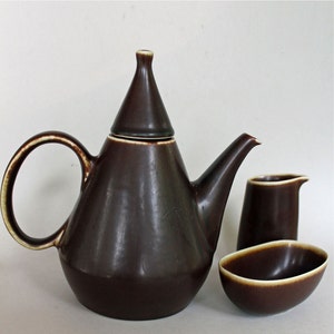 May include: A brown ceramic teapot with a cone-shaped lid, a matching creamer, and a sugar bowl. The set has a minimalist design with a light brown rim.