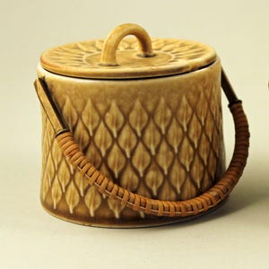Può includere: Un contenitore cilindrico in ceramica marrone con coperchio e manico intrecciato. Il contenitore presenta un motivo a losanghe e un manico curvo sul coperchio. Il manico intrecciato è realizzato in materiale marrone chiaro. Il coperchio ha un design circolare.
