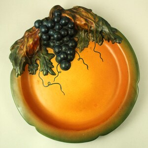 May include: A decorative, ceramic serving dish with a vibrant orange interior and a green rim. The dish is adorned with a cluster of dark purple grapes and green leaves, creating a fruit-themed design. The dish is approximately 10 inches in diameter.