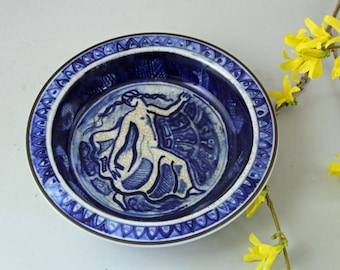 Könnte beinhalten: Eine runde, blau-weiße Keramikschale mit dekorativem Rand. Das Innere der Schale zeigt eine stilisierte weiße Figur auf blauem Hintergrund. Gelbe Blumen sind um die Schale angeordnet.