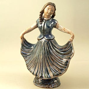 Peut inclure: Figurine en céramique d'une femme en robe fluide, les bras tendus. La robe est bleue et marron, avec des plis détaillés. La figurine a une expression sereine et est montée sur une base circulaire.