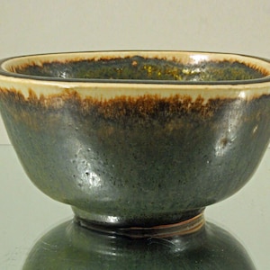 Puede incluir: Un cuenco de cerámica hecho a mano con un exterior verde oscuro y un borde color crema. El cuenco tiene una forma redondeada con una base con pie. El interior tiene un acabado brillante con toques dorados.