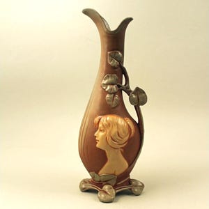 May include: A brown ceramic vase with a woman's profile in relief. The vase has a slender, elongated shape with a flared top and a decorative vine with leaves. The base is a silver color. The vase is approximately 12 inches tall.