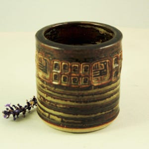 Könnte beinhalten: Eine zylindrische Keramikvase mit dunkelbrauner Außenseite und einem strukturierten, geometrischen Muster in Braun- und Goldtönen. Die Vase hat eine weite Öffnung und einen leicht ausgestellten Boden. Ein Zweig lila Blumen liegt in der Nähe der Vase.
