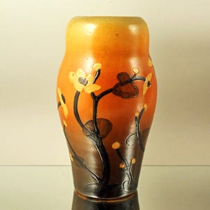 May include: A ceramic vase with a gradient of colors, transitioning from a light orange at the top to a dark brown at the base. The vase is adorned with a floral design featuring yellow flowers and dark brown stems and leaves.