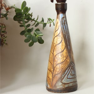 Puede incluir: Un jarrón de cerámica hecho a mano con forma cónica. El jarrón presenta una base y cuello marrones, con un cuerpo que transita de un amarillo dorado a un tono azul claro. Intrincados patrones de hojas y geométricos adornan la superficie, agregando un toque artístico.