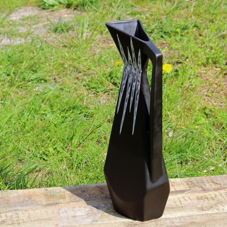 May include: A black ceramic pitcher with a geometric design. The pitcher has a wide mouth and a handle on the side. The design features white lines that create a pattern of triangles and squares.