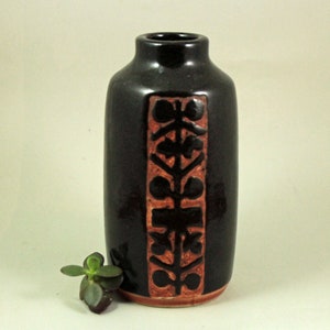 May include: A black ceramic vase with a rectangular, reddish-brown floral design. The vase has a rounded body and a narrow neck. A small green plant sits near the base of the vase.
