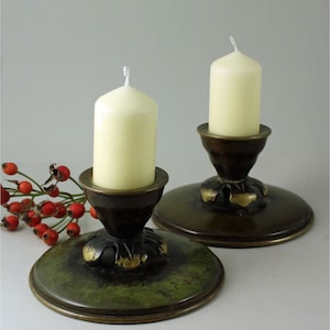 May include: Two antique brass candle holders with ivory candles. The candle holders have a dark brown base with a green and gold patina. The candles are cylindrical and have a white wick. A sprig of red berries is in the background.