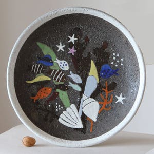 May include: A ceramic plate with a dark brown textured surface and a white rim. The plate is decorated with colorful fish, starfish, and coral. The fish are yellow, orange, blue, and white. The coral is orange. The plate is round.