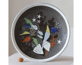 May include: A ceramic plate with a dark brown textured surface and a white rim. The plate is decorated with colorful fish, starfish, and coral. The fish are yellow, orange, blue, and white. The coral is orange. The plate is round.