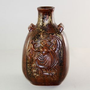 Può includere: Un vaso in ceramica marrone con un designo in rilievo di una donna che tiene una luna crescente e un uccello. Il vaso ha una smaltatura marrone macchiata e un corpo arrotondato.